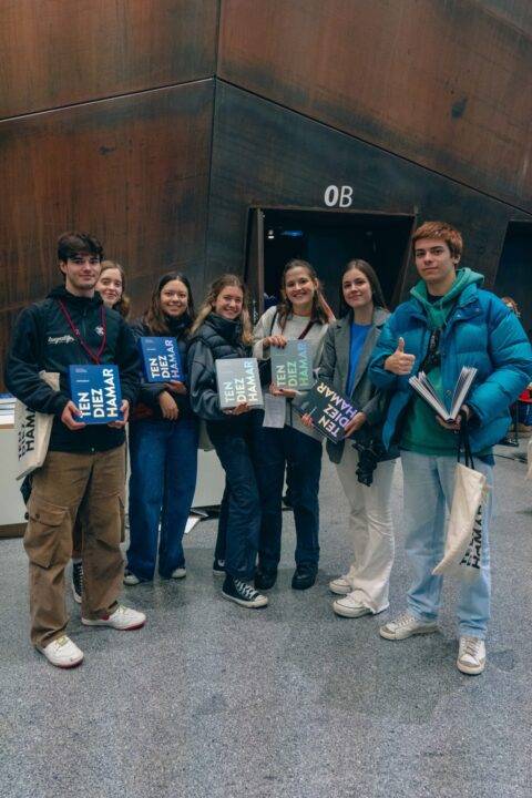 Un grupo de seis jóvenes sonrientes posan con libros en un ambiente interior moderno.