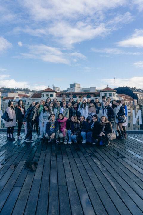 Un gran grupo de personas posa en una terraza con edificios al fondo y un cielo parcialmente nublado.