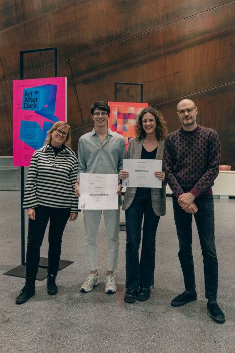Cuatro personas posan en un evento artístico, sosteniendo certificados de reconocimiento frente a obras expuestas.