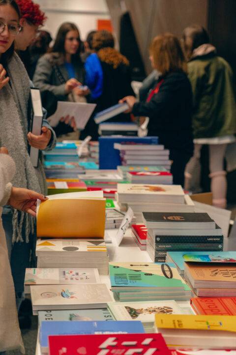 Una feria de libros donde varias personas están revisando y eligiendo libros de una larga mesa llena de publicaciones diversas.