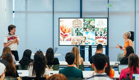 Una presentación en un aula con un expositor y una presentadora frente a una pantalla grande mostrando imágenes.