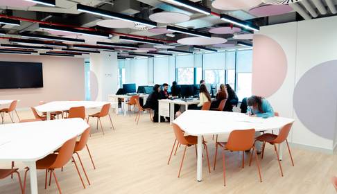 Una sala de trabajo moderna con mesas blancas y sillas naranjas, donde varias personas están colaborando.