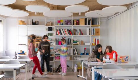 Un grupo de estudiantes interactúa en un aula moderna con estanterías llenas de libros y materiales de estudio.
