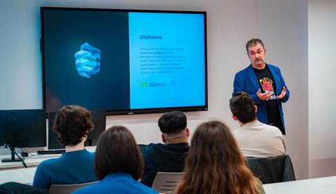 Un presentador habla frente a un grupo de personas en un aula moderna mientras se muestra una diapositiva sobre UX alliance.