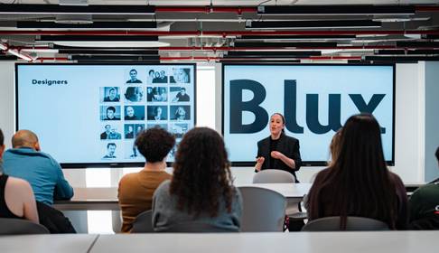 Una presentadora expone información sobre diseñadores en una sala moderna con pantallas grandes.