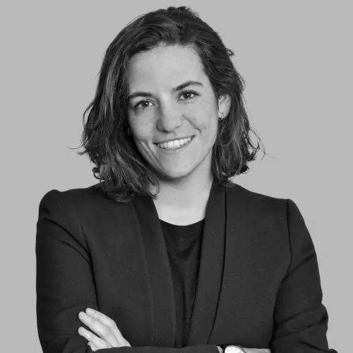 Una mujer sonriente con cabello rizado y corto, vestida con un traje negro, posa con los brazos cruzados sobre un fondo gris.