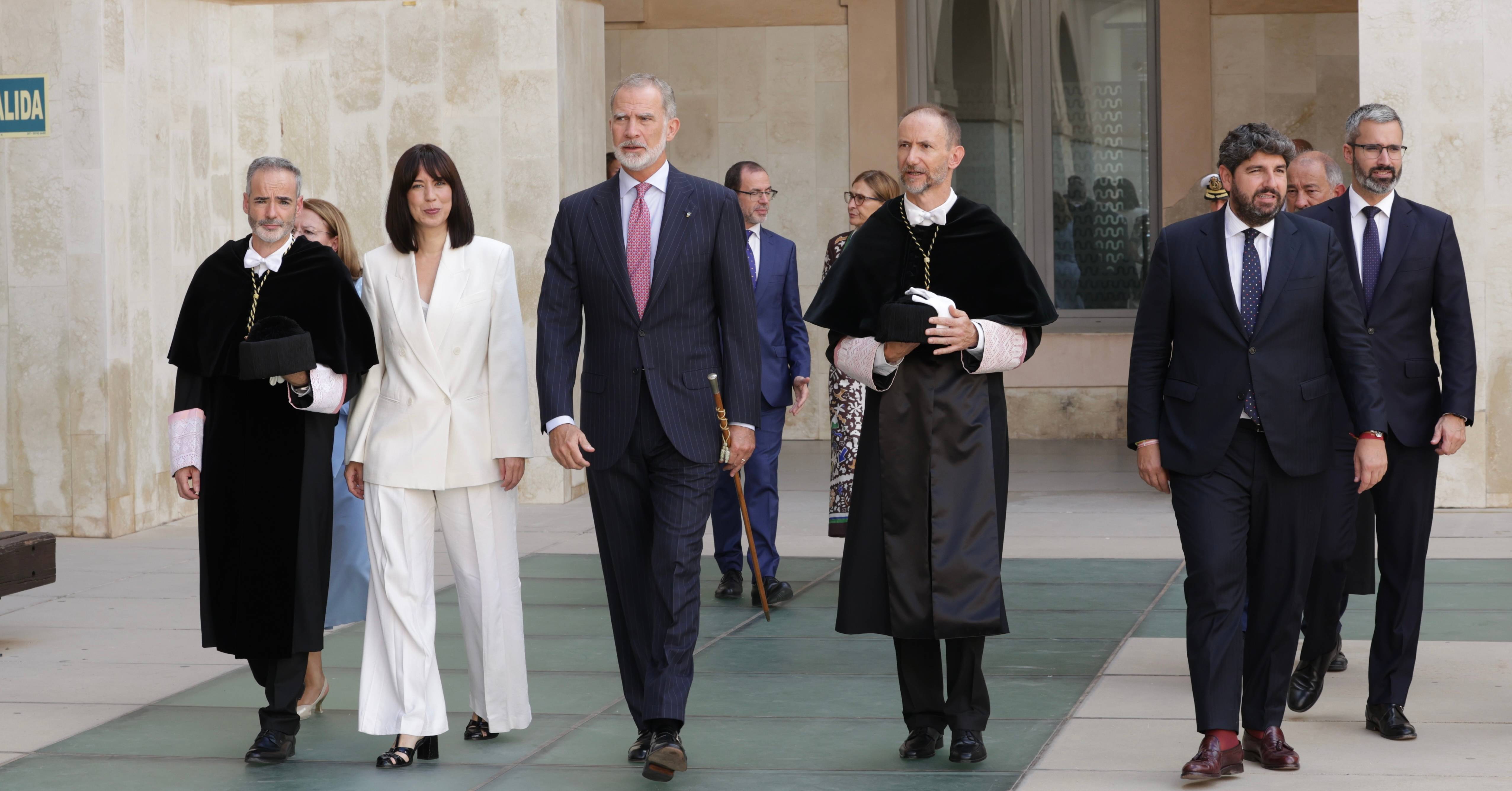 Un grupo de personas vestidas formalmente caminan hacia la cámara en un entorno arquitectónico elegante.