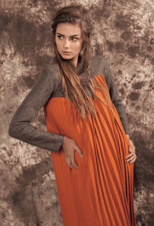 Una modelo viste un vestido naranja con pliegues, posando de forma elegante frente a un fondo texturizado.