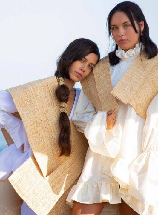 Dos mujeres posando en un entorno costero, vistiendo ropa con un diseño contemporáneo y texturas naturales.