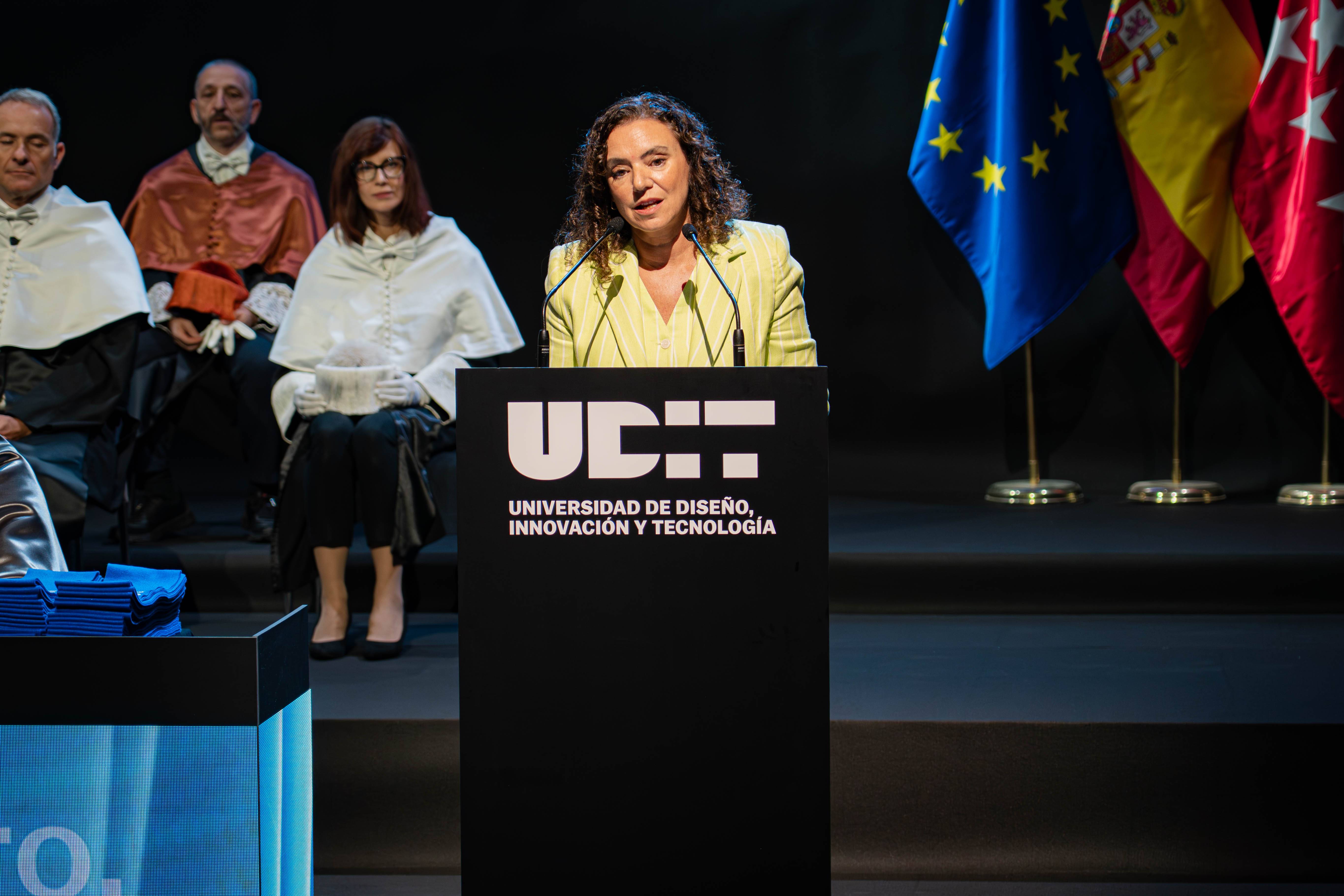 Una mujer está hablando en un evento formal frente a un podio, mientras varias personas la observan en el fondo.