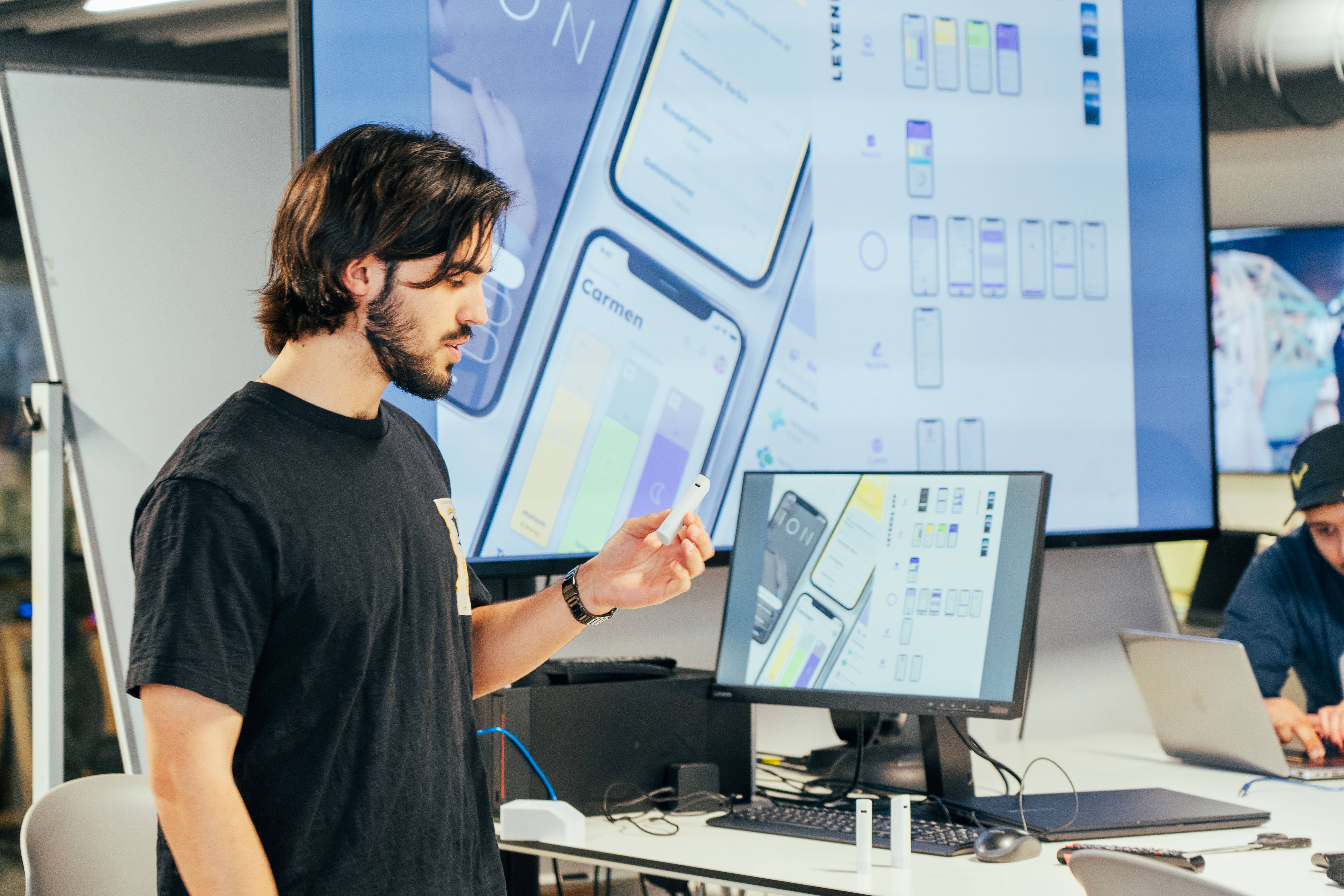 Un joven sostiene un dispositivo frente a una pantalla que muestra diseños de interfaces de usuario.