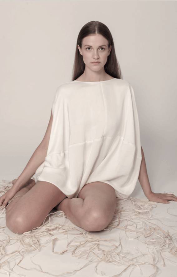 Una modelo sentada en el suelo, vistiendo una blusa blanca suelta y rodeada de hilos y elementos decorativos.
