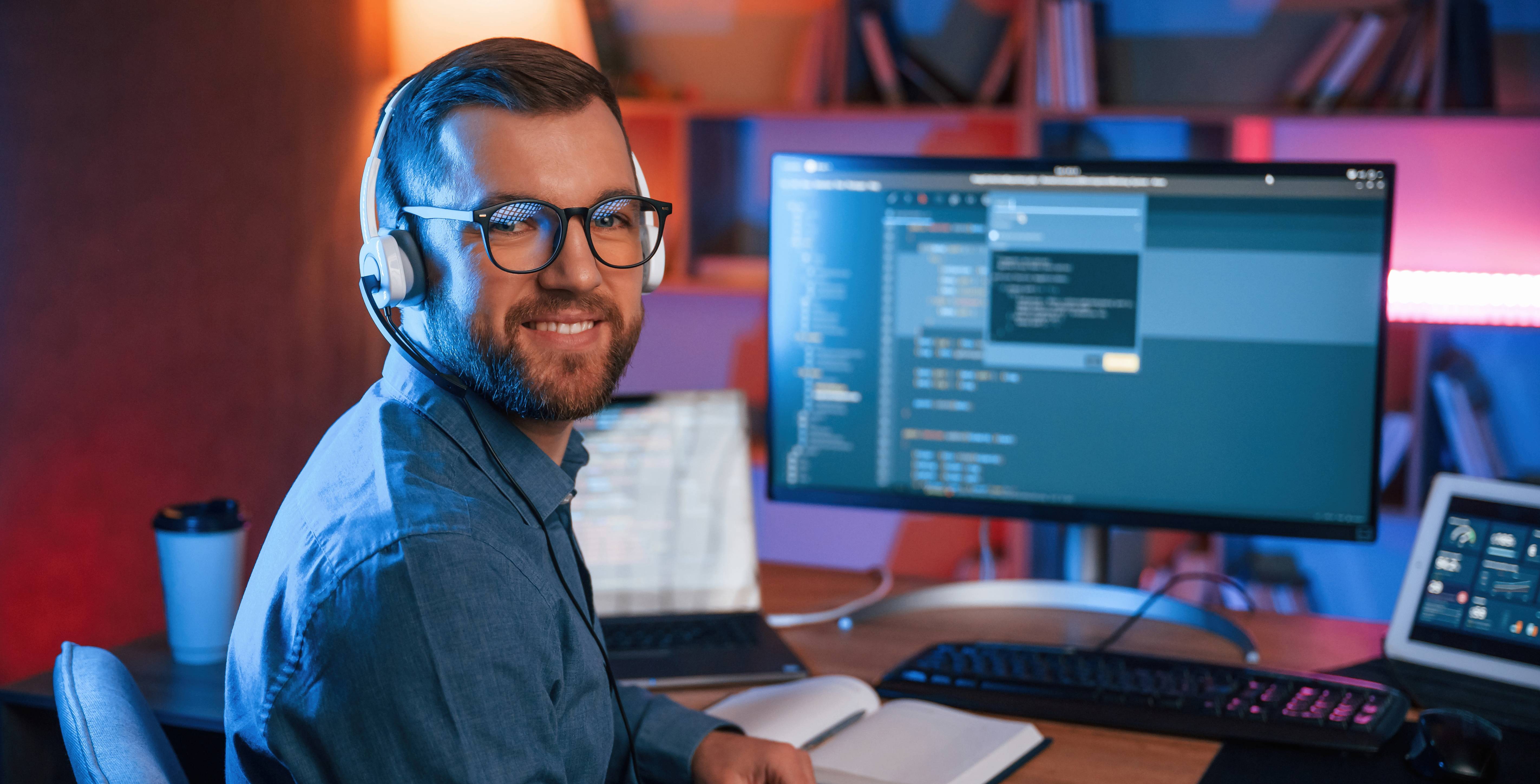 Un hombre sonriente con auriculares y gafas trabaja en una computadora en un entorno iluminado.
