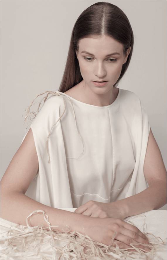 Una mujer sentada en un fondo neutro, vestida con una blusa blanca y con una expresión contemplativa.