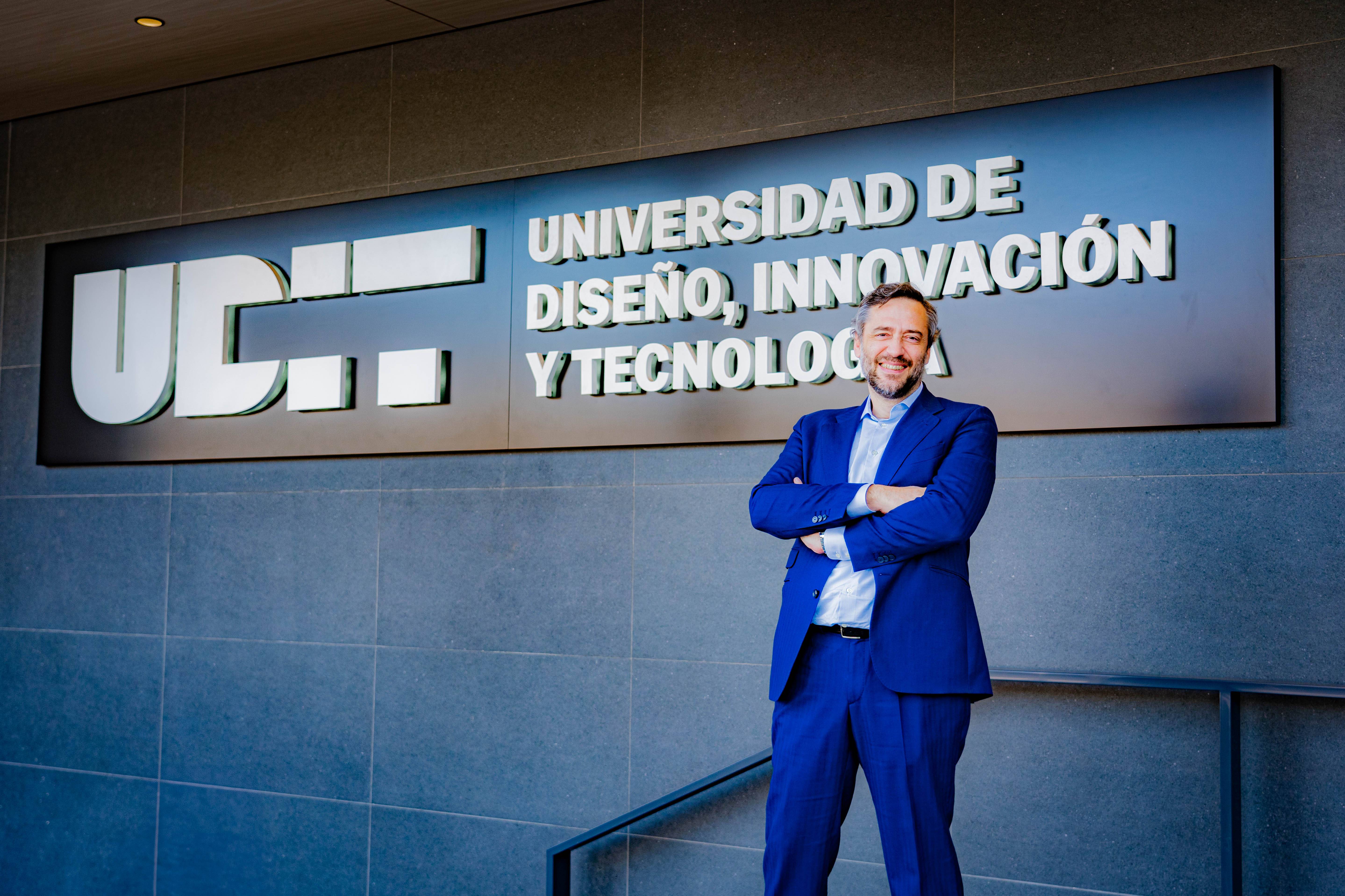 Un hombre sonriente de traje azul posa frente al letrero de una universidad que destaca su enfoque en diseño e innovación.