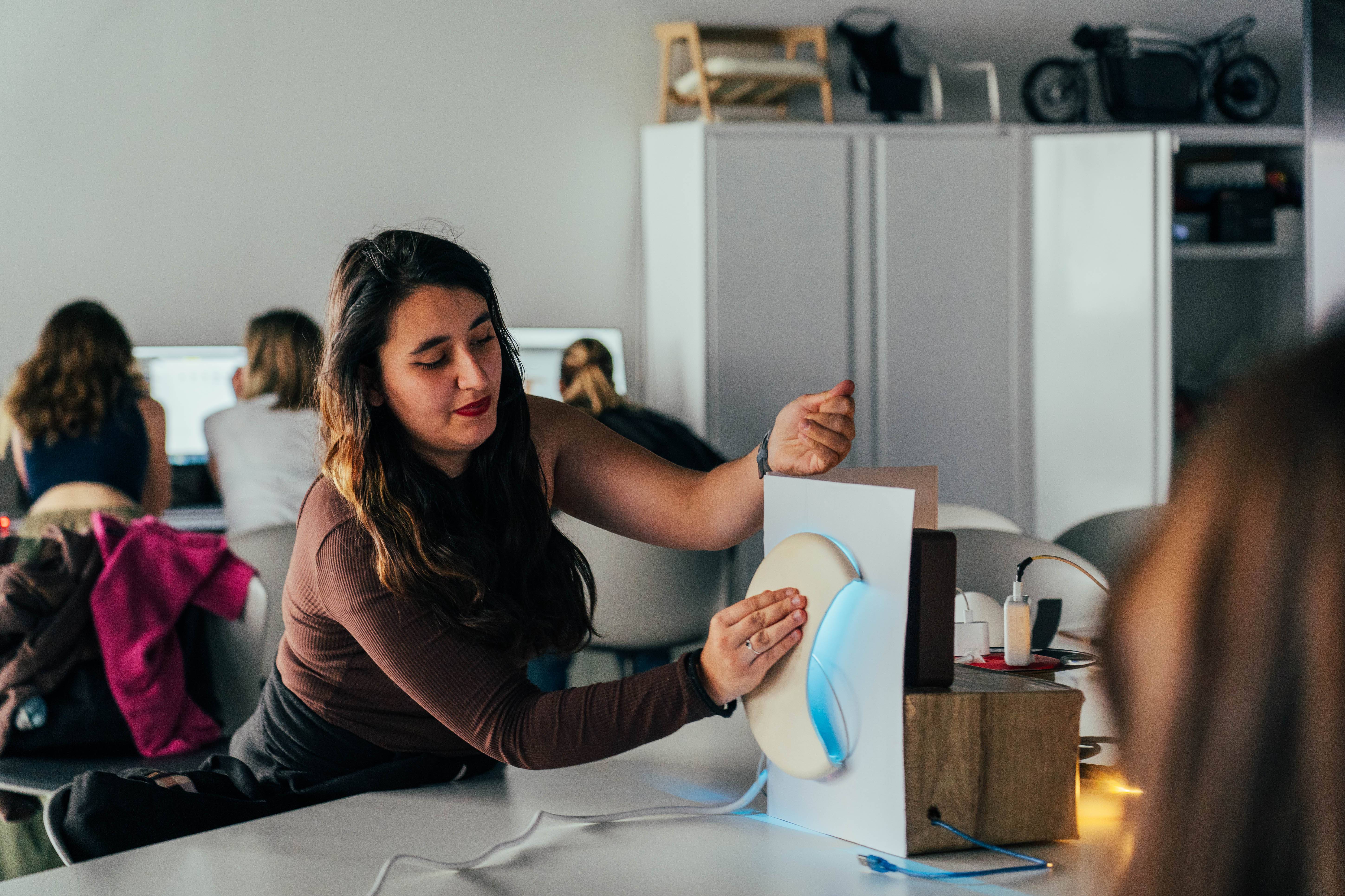 Una mujer trabaja en un proyecto mientras otros estudiantes están en sus computadoras al fondo.