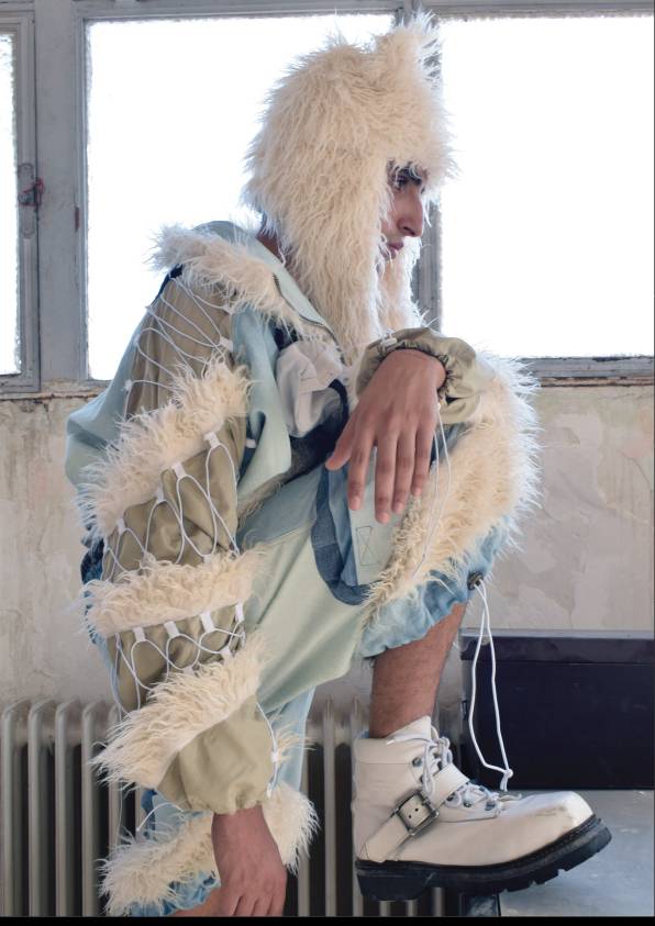 Una persona está sentada en una posición pensativa, vestida con un atuendo llamativo que incluye un gorro de pelaje y botas blancas.
