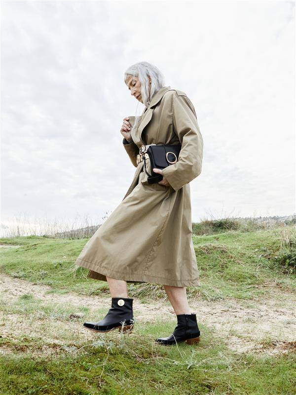 Una mujer elegante camina por un paisaje natural vistiendo un abrigo largo y botas negras.