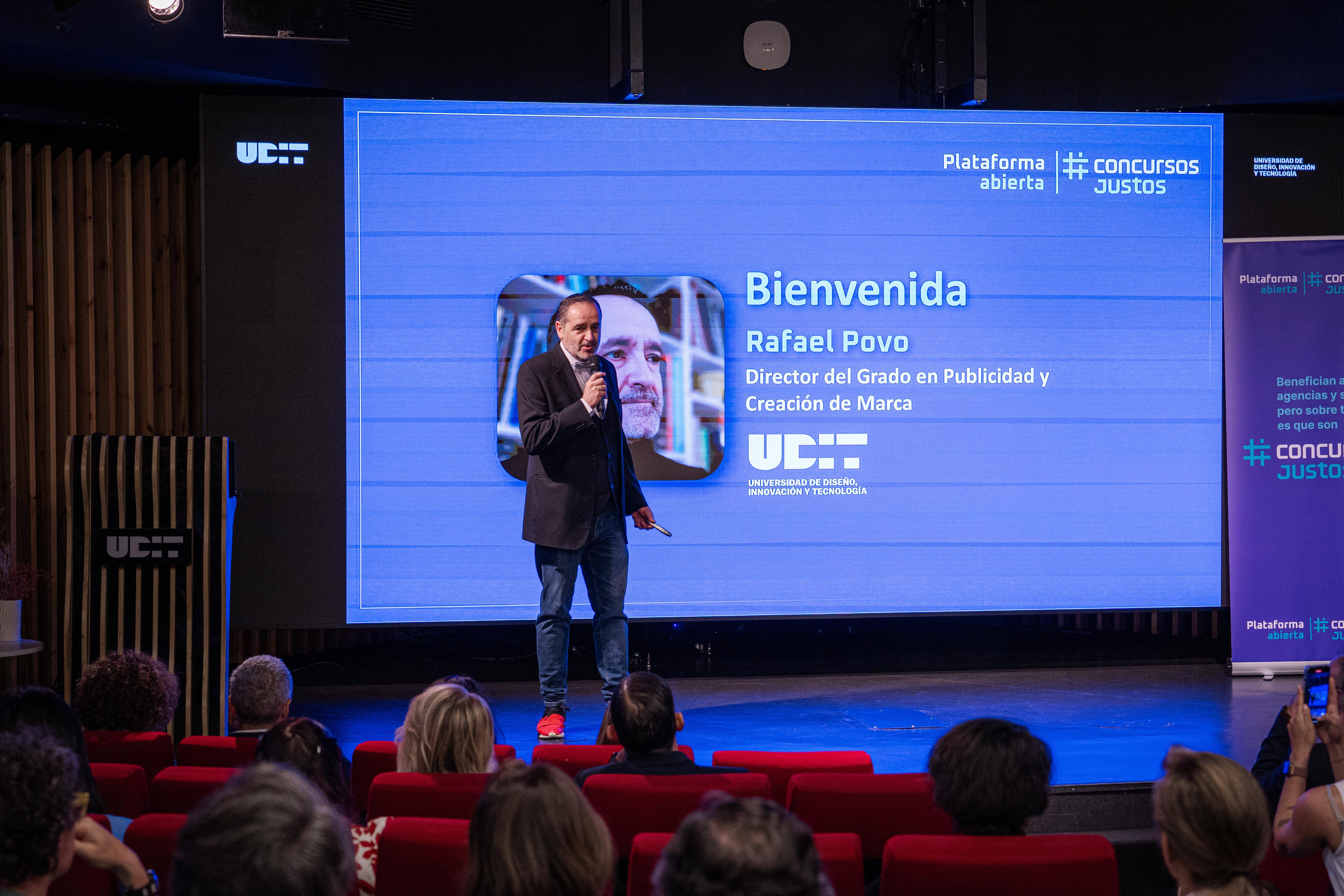 Un hombre presenta un evento en un escenario con una pantalla de fondo que muestra información respecto al mismo.