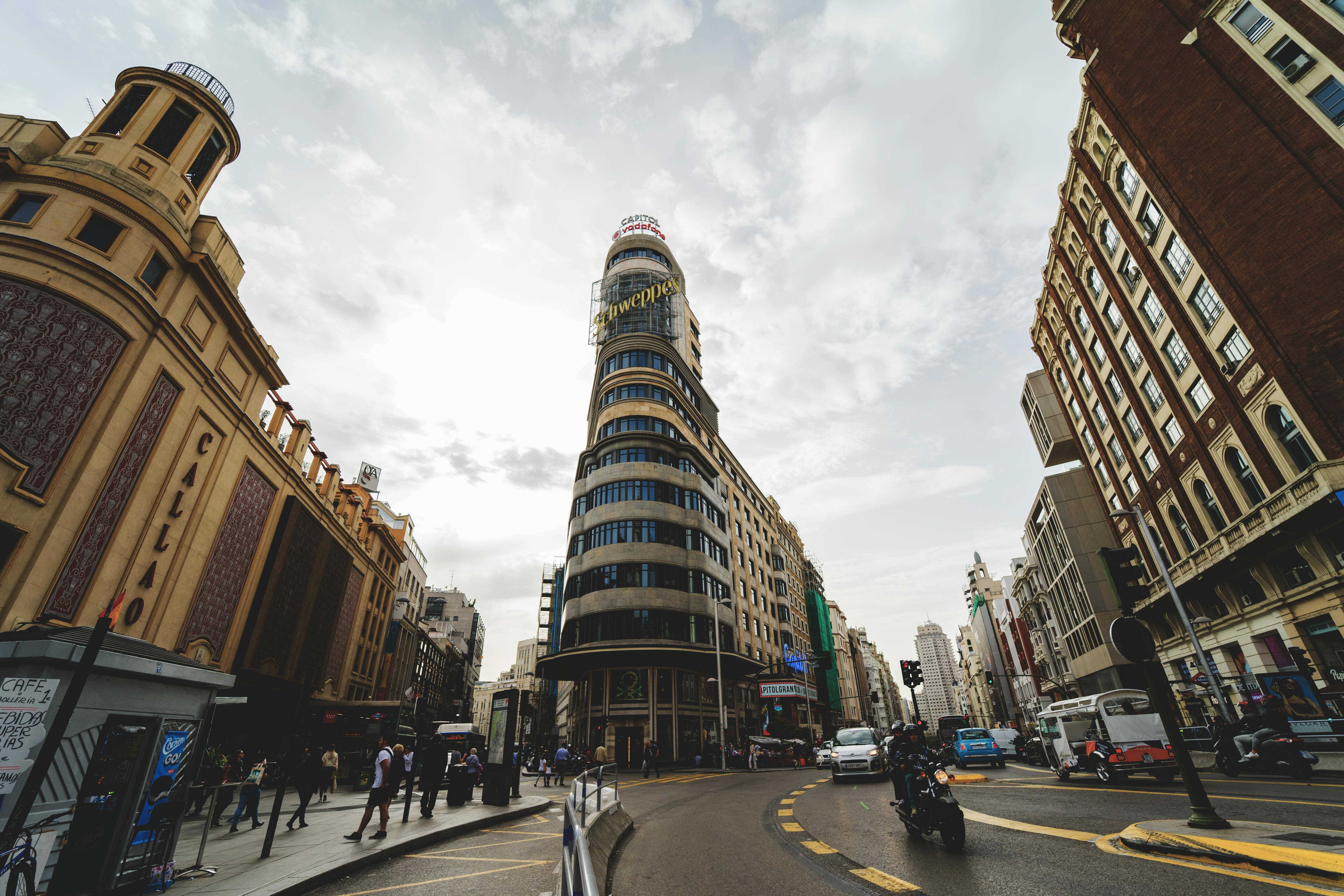 La imagen muestra una vista urbana de un alto edificio moderno en una intersección de calles con gente y vehículos.