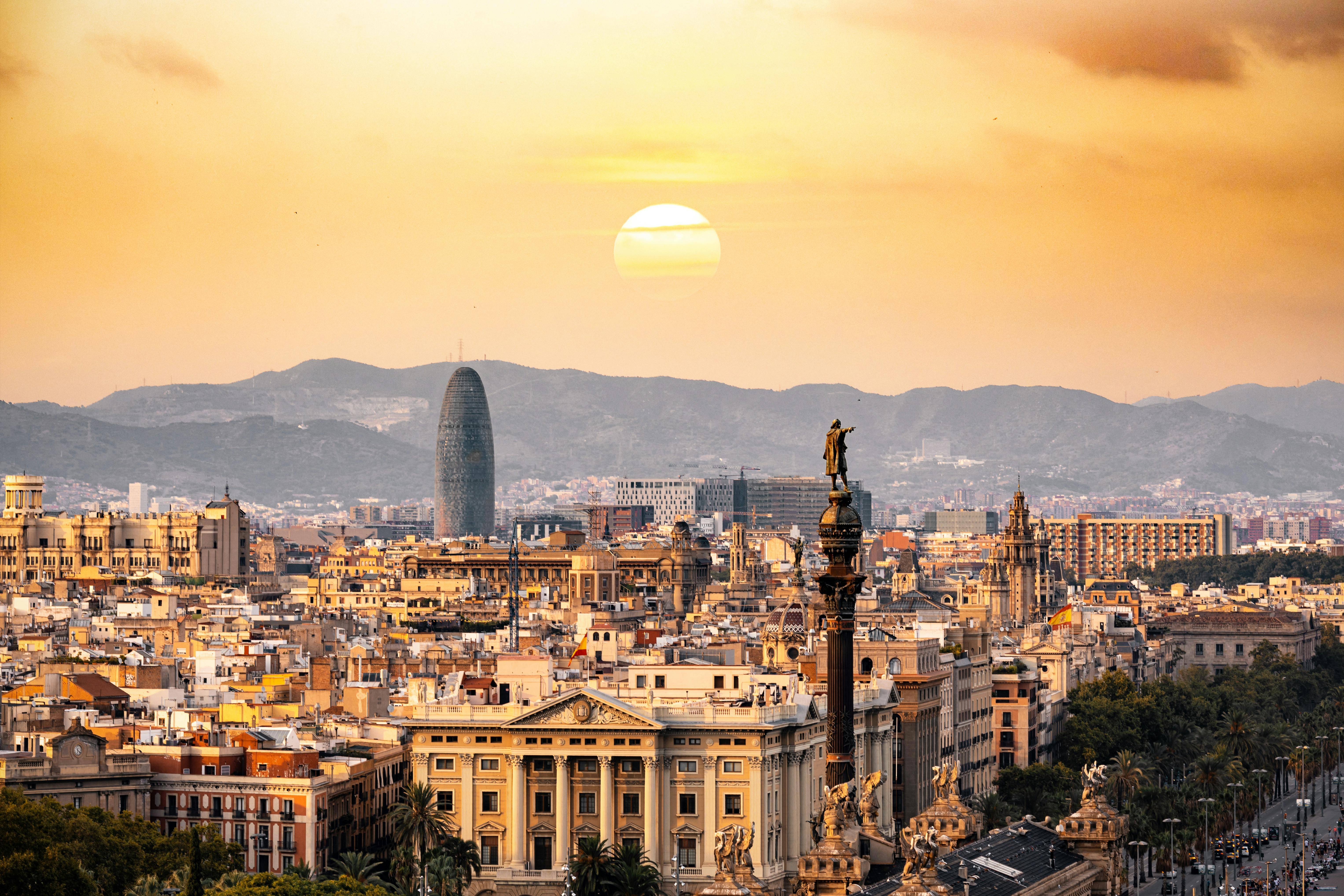 Una vista panorámica de Barcelona al atardecer con edificios emblemáticos y montañas de fondo.