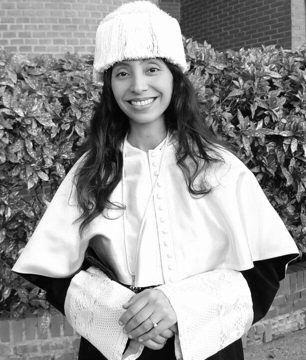 Una mujer sonriente con una toga y un gorro blanco, posando frente a un fondo de hojas verdes.