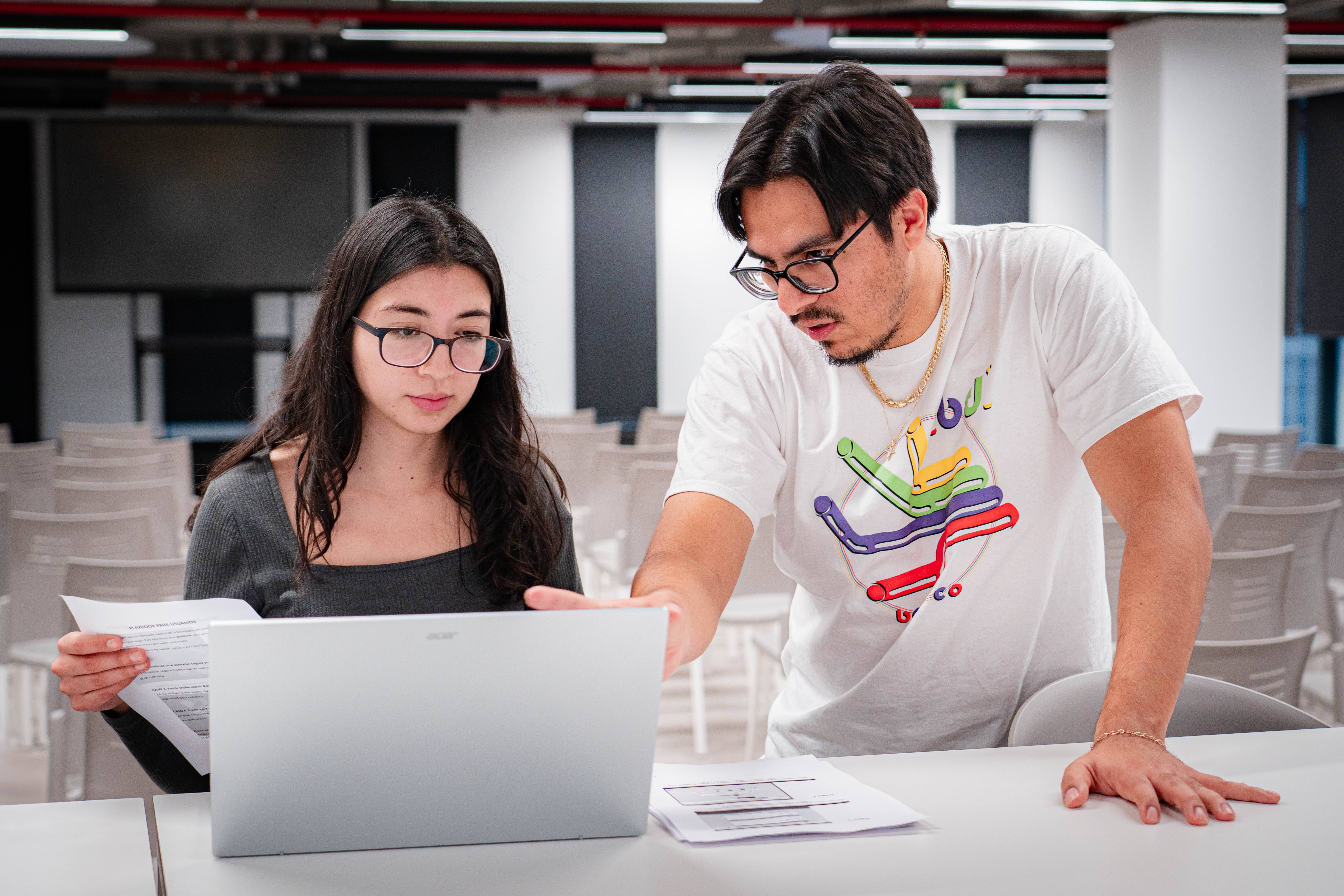 Un hombre y una mujer están colaborando en una computadora portátil mientras revisan documentos en un entorno de estudio.
