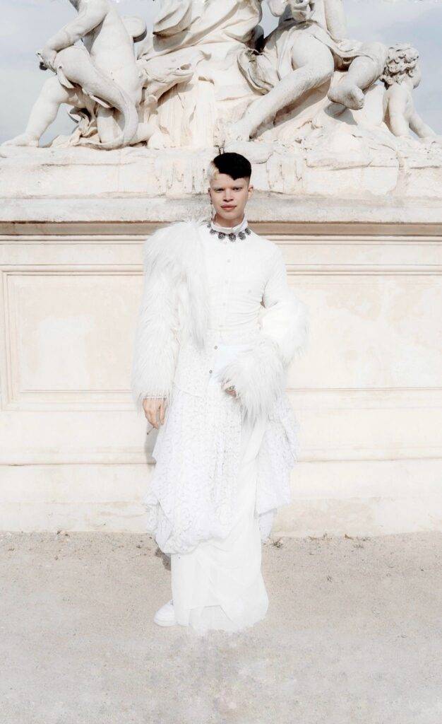 Un modelo con un vestido blanco y detalles de plumas posa frente a una escultura clásica.