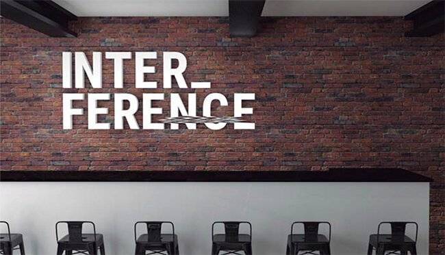 Una pared de ladrillo con la palabra 'INTERFERENCE' escrita en letras blancas, sobre un bar con sillas negras.