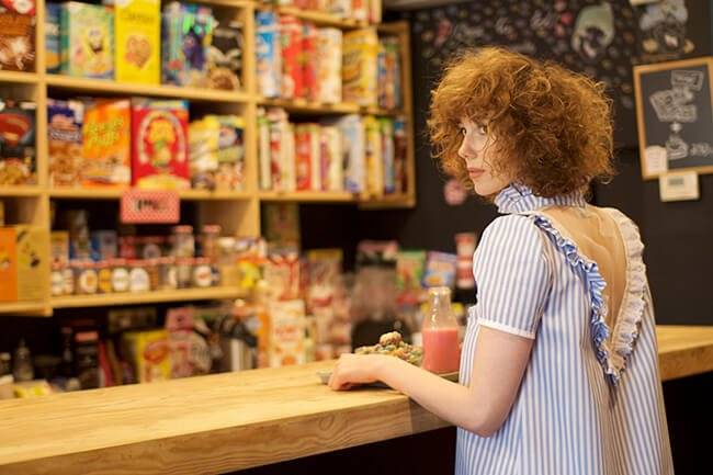 Una joven con cabello rizado observa desde el mostrador de una tienda llena de cereales coloridos en el fondo.