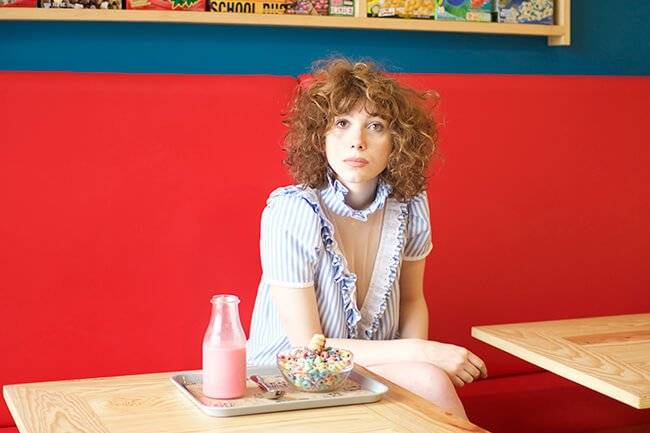 Una persona joven sentada en un banco de una cafetería, con una botella de bebida y un plato de cereales frente a ella.