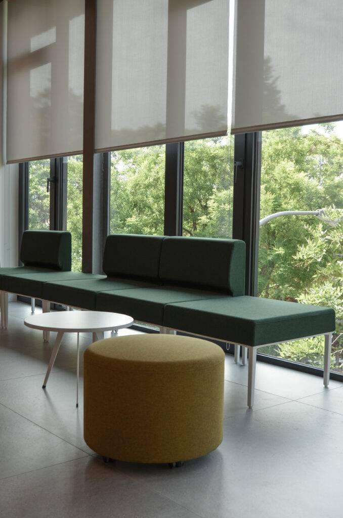 Una sala moderna con un sofá verde, una mesa blanca y un taburete amarillo, rodeada de grandes ventanales que dan a un paisaje verde.