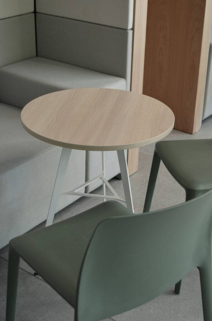 Una mesa redonda de madera clara junto a dos sillas verdes en un ambiente moderno.