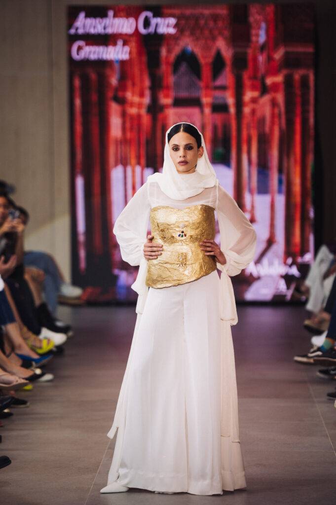 Modelo desfilando con un atuendo elegante y un abrigo dorado en un evento de moda.