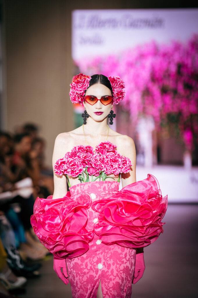 Una modelo viste un vestido rosa decorado con flores, luciendo un estilo vibrante y audaz en una pasarela.