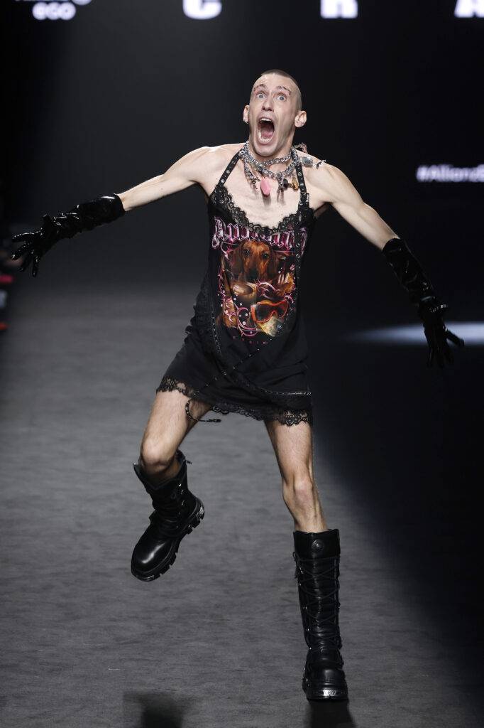Un modelo masculino luciendo un vestido negro con detalles y botas altas, en una pasarela con una expresión intensa.