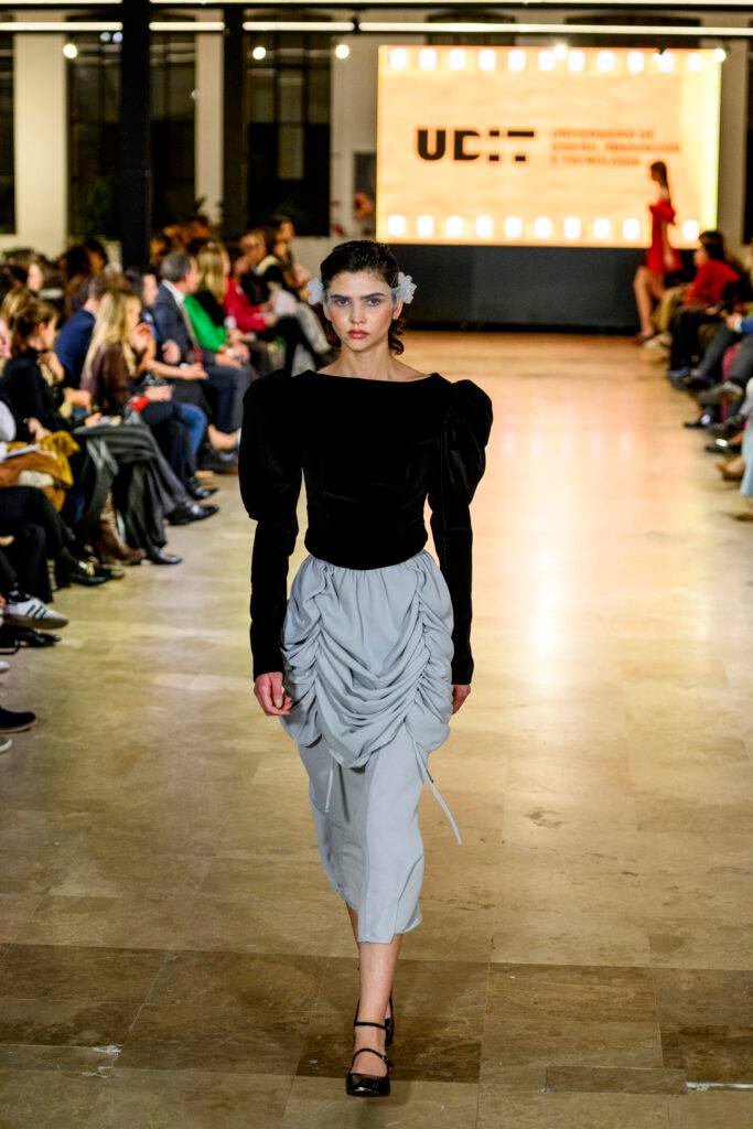 Una modelo camina por la pasarela durante un desfile de moda con un vestido negro y una falda gris.