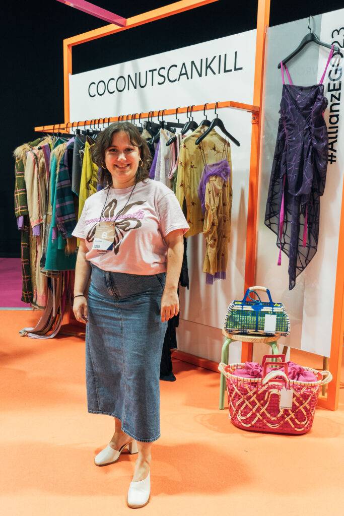 Una mujer sonriente posa frente a un fondo con ropa expuesta y un letrero que dice COCONUTSCANKILL.
