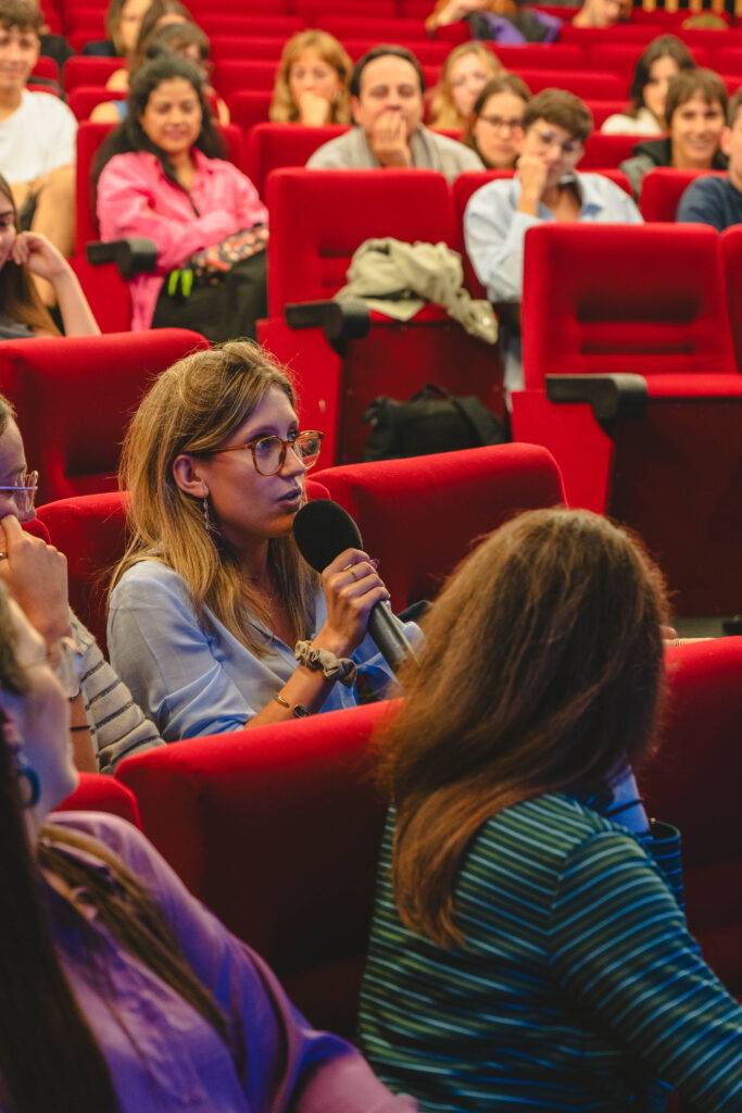 Una mujer con gafas levanta la mano para hablar en una sala llena de público sentado en asientos rojos.