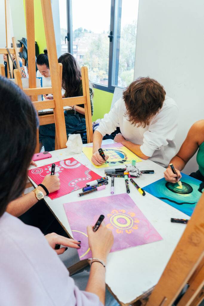 Un grupo de personas en una sala iluminada trabajando en proyectos artísticos con marcadores.