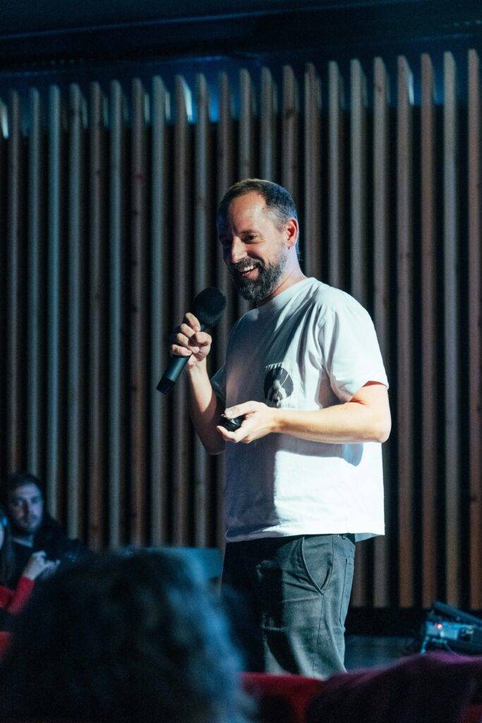 Un hombre con barba presenta en un escenario mientras sostiene un micrófono.