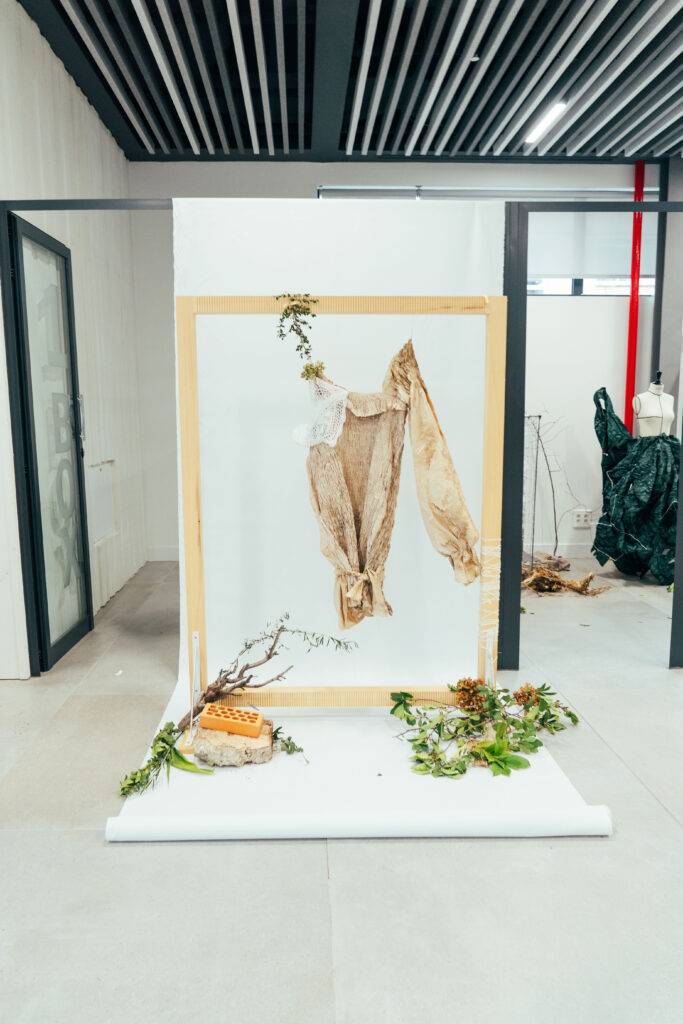 Un marco de madera sostiene una prenda de ropa suspendida, rodeada de decoraciones naturales en un ambiente artístico.