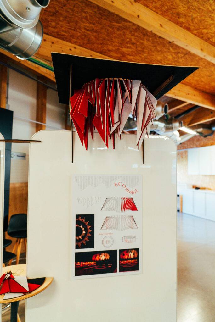 La imagen muestra una exhibición de arte con estructuras de papel en rojo y blanco colgando del techo, y un panel informativo en la parte inferior.
