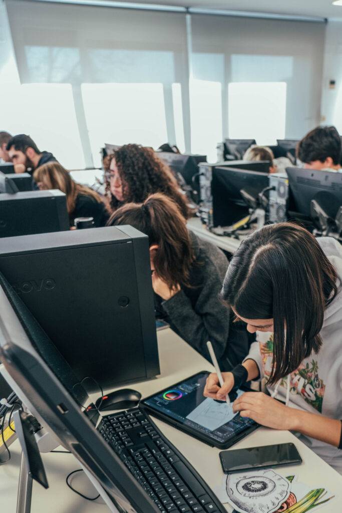 Un grupo de estudiantes trabaja en computadoras mientras una persona dibuja en una tablet.
