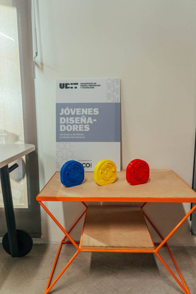 Tres objetos decorativos de colores sobre una mesa junto a un cartel que dice 'JÓVENES DISEÑADORES'.