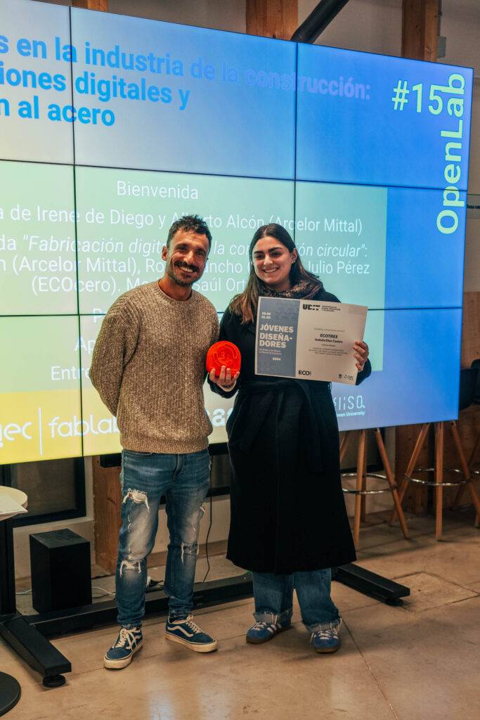 Dos personas posan sonrientes frente a una pantalla que presenta un evento relacionado con la industria de la construcción.