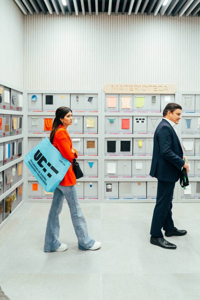 Una mujer con un abrigo rojo y un bolso azul camina junto a un hombre de traje en un espacio moderno con exhibiciones de materiales.