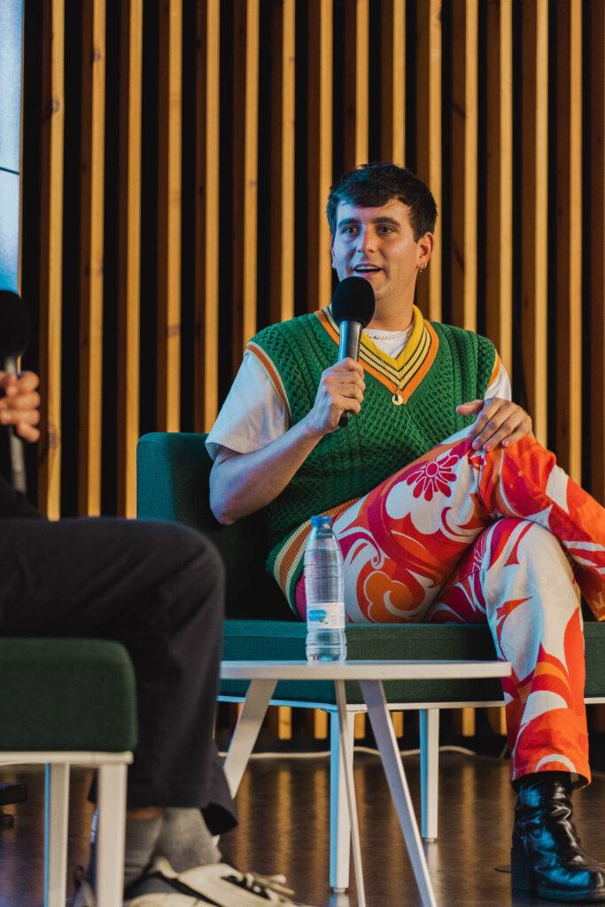 Un joven vestido con un suéter colorido y pantalones llamativos habla en un evento.