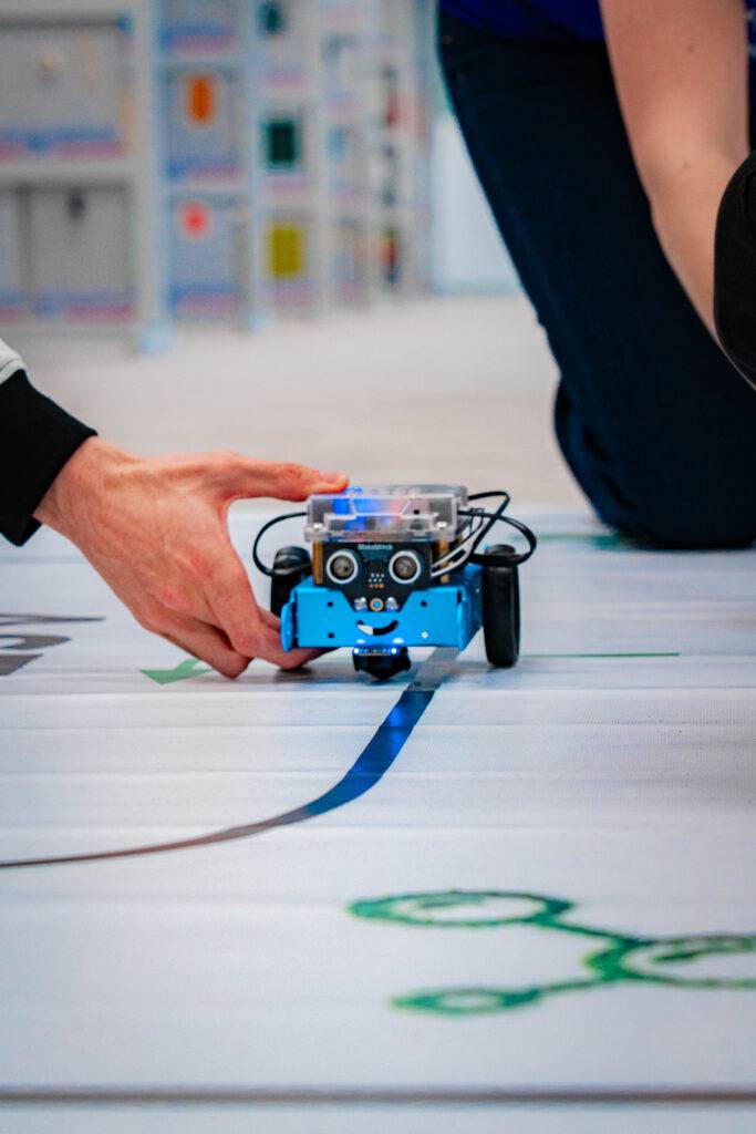 Un robot azul sigue una línea en un suelo decorado mientras una persona lo guía con la mano.