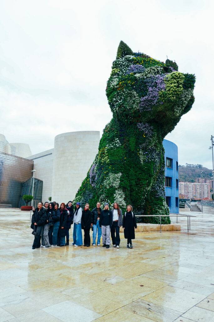 Un grupo de personas posa frente a una enorme escultura de un perro hecha de plantas en un entorno urbano.